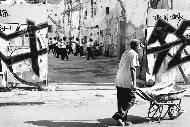 Black and white image of exercise group taken from the street. A man with a wheelbarrow is to the right of the shot.